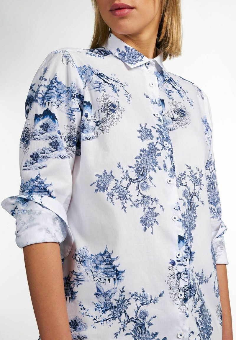 Femme portant une chemise blanche à boutons avec un imprimé floral bleu et pagode, manches retroussées, et rouge à lèvres rose clair.