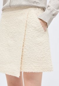 Γυναίκα που φοράει υφασμάτινη, εκρού φούστα τύπου wrap με φθαρμένες άκρες, με το χέρι στην τσέπη, συνδυασμένη με ανοιχτό γκρι μακρυμάνικη μπλούζα.