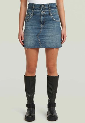 Kobieta w średnio niebieskiej dżinsowej mini spódnicy z guzikami z przodu oraz wysokich czarnych skórzanych butach stoi na szarym tle.