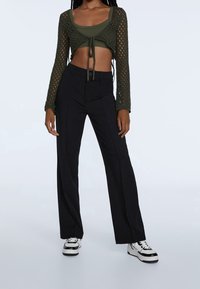 Olijfgroene, lange mouw, opengehaakte crop top met strikdetail, gecombineerd met zwarte, wijde broek en witte sneakers met zwarte accenten.