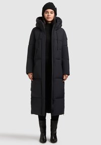 Cappotto imbottito lungo e nero con cappuccio, zip frontale e due tasche laterali. Indossato sopra un completo nero, abbinato a stivali neri fino al ginocchio.