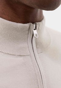 Lys beige sweater med en høj ribbet krave, der har en halvlång lynlås med en sølvfarvet trækklap. Fin strikket tekstur.