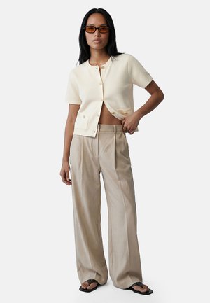 Mujer con pantalones beige de pata ancha, cárdigan corto de manga corta color crema abotonado, gafas de sol y sandalias negras con puntera cuadrada, posando con la mano en la cintura.