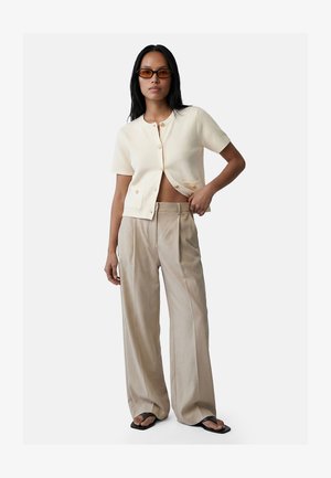 Kvinde iført beige bukser med vide ben, cremefarvet kortærmet cardigan med knapper, solbriller og sorte sandaler med firkantet tå, poserer med hånden på hoften.