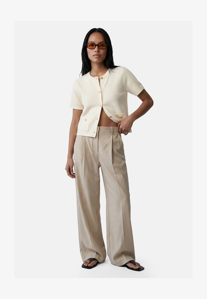 Woman wearing beige wide-leg trousers, cream buttoned short-sleeve cardigan, sunglasses, and black square-toe sandals, posing with hand on waist.