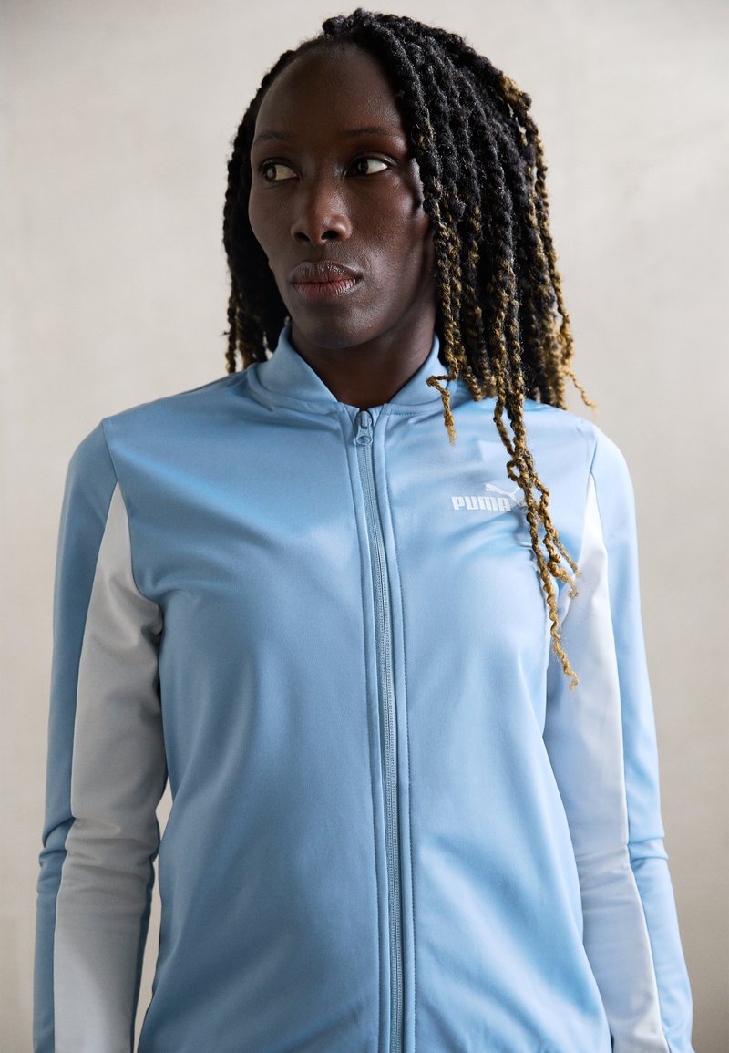 Svetlomodrá športová bunda na zips s šedými rukávmi, vyrobená z hladkej látky. Má vysoký golier a nenápadné logo na ľavej hrudi.