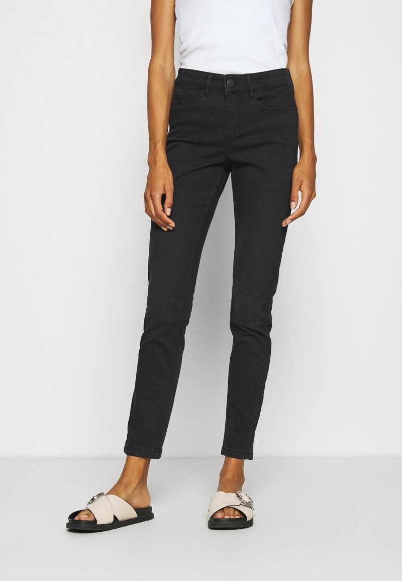 Jean skinny en denim noir avec une taille mi-haute, cinq poches et une texture lisse, associé à des sandales plates de couleur claire.