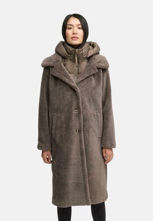 Manteau marron, fluffy et surdimensionné avec un col, des poches latérales et deux boutons. En dessous, une veste matelassée sombre avec un col montant.