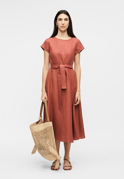 Γυναίκα φοράει ρουμπινί midi φόρεμα με ζώνη και καφέ σανδάλια, κρατώντας μεγάλη πλεκτή τσάντα ώμου με ταμπά χερούλια.