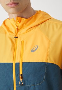 Chaqueta con capucha de color naranja brillante en la parte superior y paneles inferiores en color teal. Cuenta con un cierre frontal con cremallera, tela texturizada y un logo en el pecho.