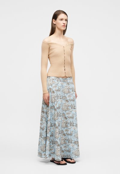 Γυναίκα που στέκεται φορώντας μια μπεζ μπλούζα με μακριά μανίκια και κουμπιά που αφήνει ακάλυπτους τους ώμους και μια μπλε φούστα μάξι με σχέδια, συνδυασμένη με μαύρα σανδάλια.