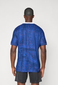 Tricou sportiv cu un model albastru și roșu, cu guler alb și mâneci scurte, având un material texturat și accente contrastante la mâneci.