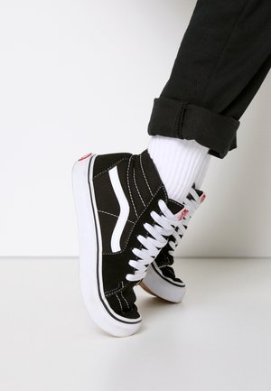 Sort-hvide høje sneakers med hvide snørebånd båret med hvide sokker og sorte opsmøgede bukser på en hvid gulv og baggrund.
