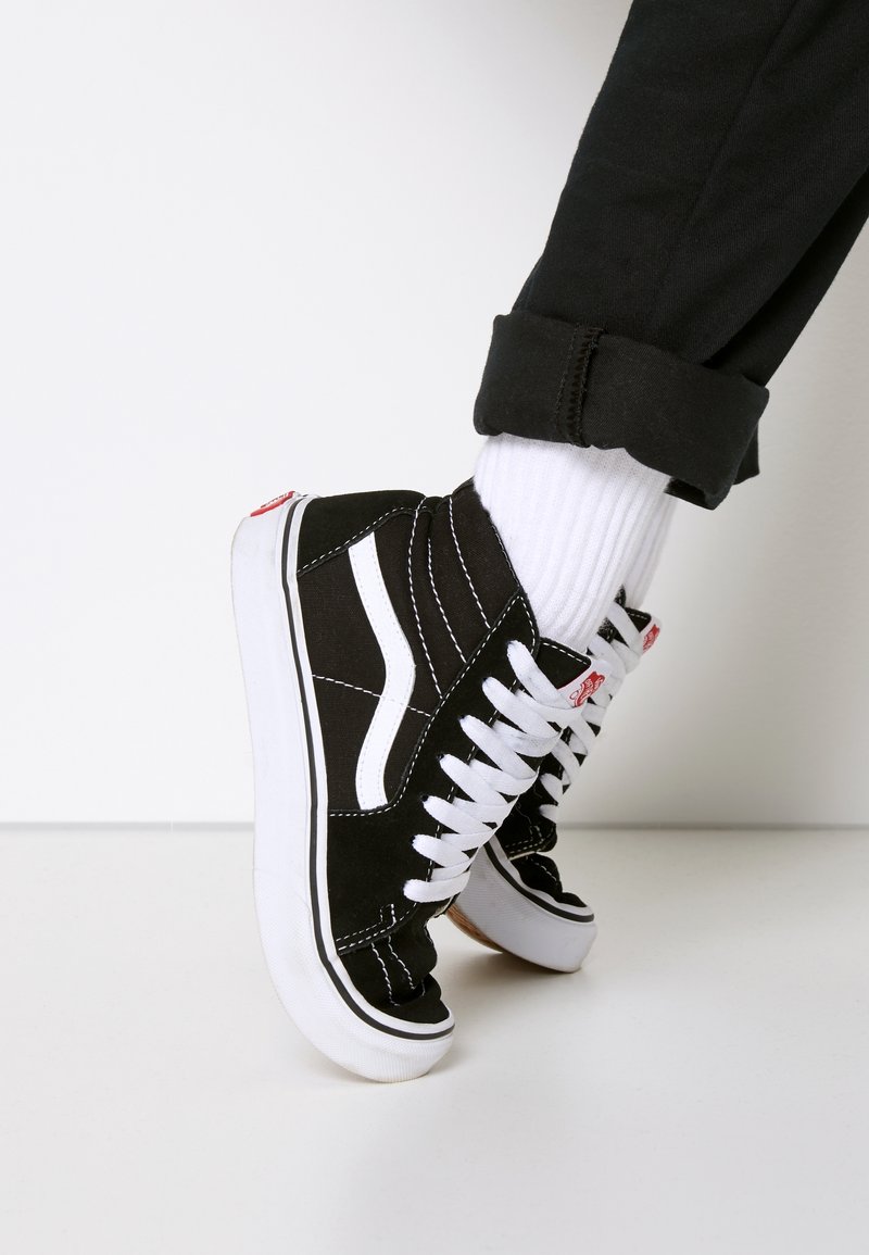 Baskets montantes noires et blanches avec des lacets blancs, portées avec des chaussettes blanches et un pantalon noir retroussé sur un sol et un fond blancs.