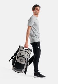 Grauer und schwarzer Sport-Rucksack mit Mesh-Seitentaschen, mehreren Reißverschlüssen und Logoakzenten. Das Modell trägt ein graues Shirt und schwarze Hosen.