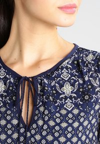 Mörkblå blus med blommiga och geometriska mönster, nyckelhålsutskärning och snörningar. Mjuk tyg med gathers på axlarna.