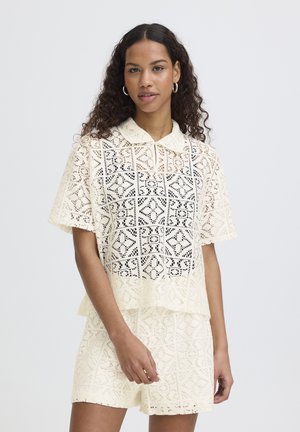 Vrouw draagt een crèmekleurige korte mouw top van gehaakte kant met kraag en bijpassende korte broek, staand tegen een effen lichte achtergrond.