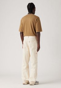 T-shirt in cotone marrone, vestibilità comoda, abbinato a pantaloni cargo color crema con tasche laterali e accento del logo rosso vicino alla tasca posteriore.