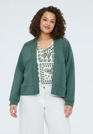 Kobieta o kręconych włosach, ubrana w zielony kardigan z rozpiętym przodem, białe spodnie i białą bluzkę z wzorem oraz dekoltem w serek, stojąca na jednolitym tle.