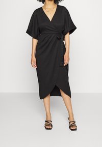 Svart omslagsklänning med V-ringning och korta ärmar, med ett knytband i midjan och en knä-lång kjol med asymmetrisk fåll. Slät tygtextur.