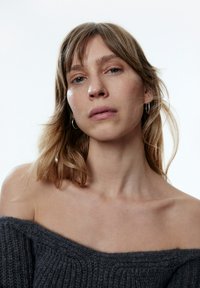 Grauer, gestrickter Pullover mit schulterfreiem Design, gerippter Textur und weitem Ausschnitt. Das Model hat hellbraune Haare und kleine Creolen.