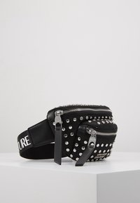 Černá ledvinka vyrobená z umělého materiálu, zdobená stříbrnými hroty, dvojitými zipy a texturovaným popruhem s bílým nápisem. Kompaktní design.