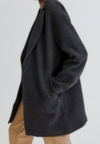 Manteau en laine rayé gris et noir, manches longues, col châle, fermeture à un seul bouton, associé à un pantalon beige.