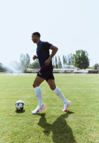 Puma nogometni igrač nosi tamnoplavi dres, crne kratke hlače i bijele čarape s ružičastim akcentima, trči po travi s nogometnom loptom.