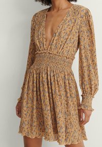Vestido amarillo mostaza con un profundo escote en V, mangas largas, cintura fruncida y estampado floral en azul y púrpura. Tejido texturizado con detalles de volantes.