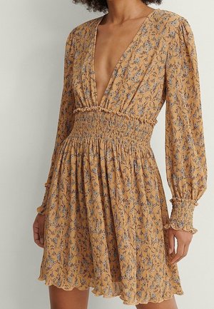 Mosterdgele jurk met een diepe V-hals, lange mouwen, gerimpelde taille en bloemenprint in blauw en paars. Getextureerde stof met gerimpelde accenten.