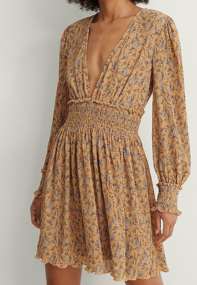 Vestido amarillo mostaza con un profundo escote en V, mangas largas, cintura fruncida y estampado floral en azul y púrpura. Tejido texturizado con detalles de volantes.