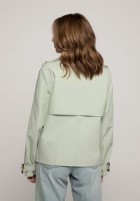 Lichtgroene jas met een gestructureerd ontwerp, voorzien van een achterkant met flapdetail en opgerolde mouwen. Gecombineerd met lichtblauwe denim broek.
