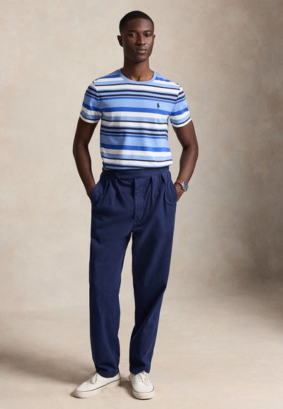 Homme debout les mains dans les poches, portant un t-shirt rayé bleu et blanc, un pantalon plissé bleu marine, des baskets blanches et une montre-bracelet.