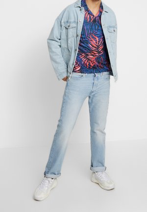 Lichtblauwe spijkerjas over een donker, kleurrijk bloemmotief overhemd, gecombineerd met lichtblauwe spijkerbroeken en witte sneakers.