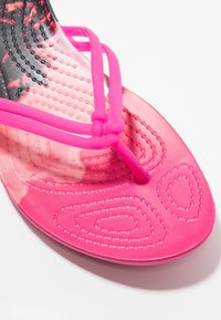 Rosa gummitong-sandal med texturerad sula och dubbla remmar som bildar en knut nära tån, på en vit bakgrund.