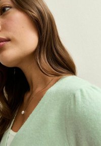 Cárdigan de punto suave en verde claro, con escote en V y hombros abullonados. Acompañado de una cadena dorada con un colgante de perla.