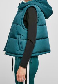 Tyrkysová, bezrukává péřová vesta s kapucí, která má přední zip a boční kapsy. Hladký materiál a nastavitelné šňůrky ve spodní části.