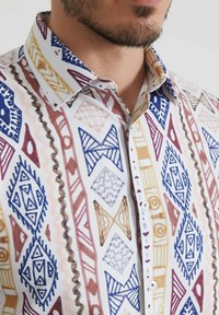 Chemise blanche à boutons avec un motif géométrique en bleu, marron et doré. Présente un col évasé et un tissu texturé avec divers motifs.