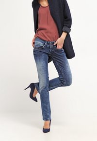 Kvinna klädd i blå skinny jeans, rostfärgad topp, marinblå strukturtröja och marinblå högklackade skor, poserar med ett ben lyft.