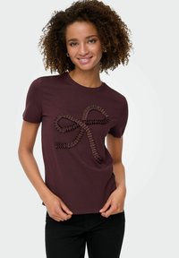 Młoda kobieta z kręconymi włosami, nosząca ciemnofioletowy t-shirt z teksturowanym wzorem kokardy, w połączeniu z czarnymi spodniami, uśmiechająca się do aparatu.