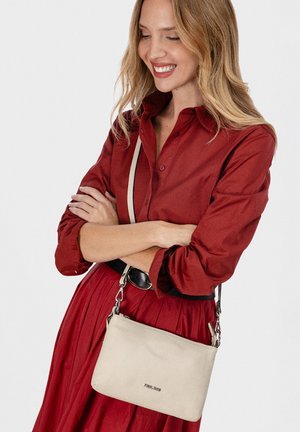 Mujer sonriente con vestido rojo y cinturón negro, brazos cruzados, llevando un bolso cruzado beige con herrajes plateados sobre fondo blanco.