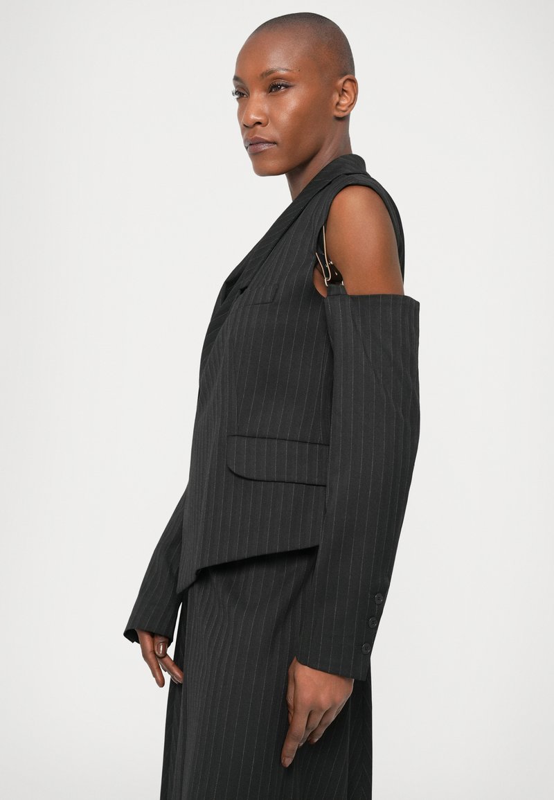 Schwarzer Pinstripe-Blazer mit einem einzigartigen asymmetrischen Design, einem abgenommenen Ärmel und einem goldenen Hardware-Detail. Glatte Textur, taillierte Passform.