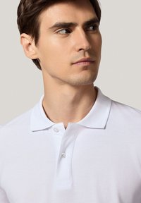 Biała bawełniana koszulka polo z kołnierzykiem zapinanym na trzy guziki, krótkimi rękawami i teksturowanym materiałem, noszona przez modela zwróconego lekko w bok.