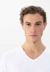 Witte v-hals t-shirt met korte mouwen, gemaakt van gladde stof. Model heeft kort, golvend haar en een subtiele oorbel.