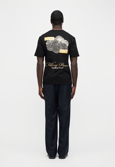 Άνδρας που φοράει μαύρο T-shirt με λευκό μοτίβο που μοιάζει με δίχτυ και κίτρινο κείμενο "Filling Pieces 'Breaking Bread'" στην πλάτη, συνδυασμένο με σκούρο παντελόνι με ρίγες.