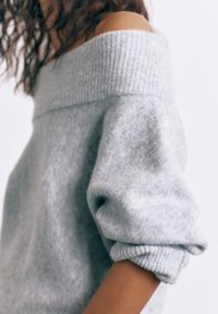 Žena nosí světle šedý svetr přes ramena s vyhrnutými rukávy na jednobarevném bílém pozadí.