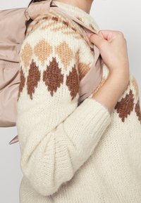 Pull crème tricoté avec des motifs en losange marron ; le modèle tient une sangle de sac à dos beige, mettant en valeur la texture et la forme.