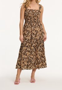 Vestido de estampado de leopardo en tonos marrón y beige con corpiño fruncido, tirantes con volantes y falda fluida de longitud midi. Lleva sandalias rosas con tacón ancho.