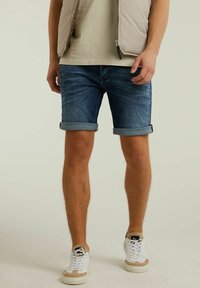 Shorts de mezclilla en azul oscuro, con dobladillos enrollados; combinados con zapatillas blancas y beiges. El modelo lleva un chaleco beige claro y una camiseta debajo.