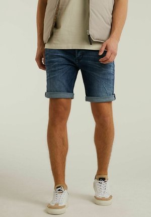 Shorts en denim bleu foncé, avec des revers ; associés à des baskets blanches et beiges. Le modèle porte un gilet beige clair et un haut en dessous.